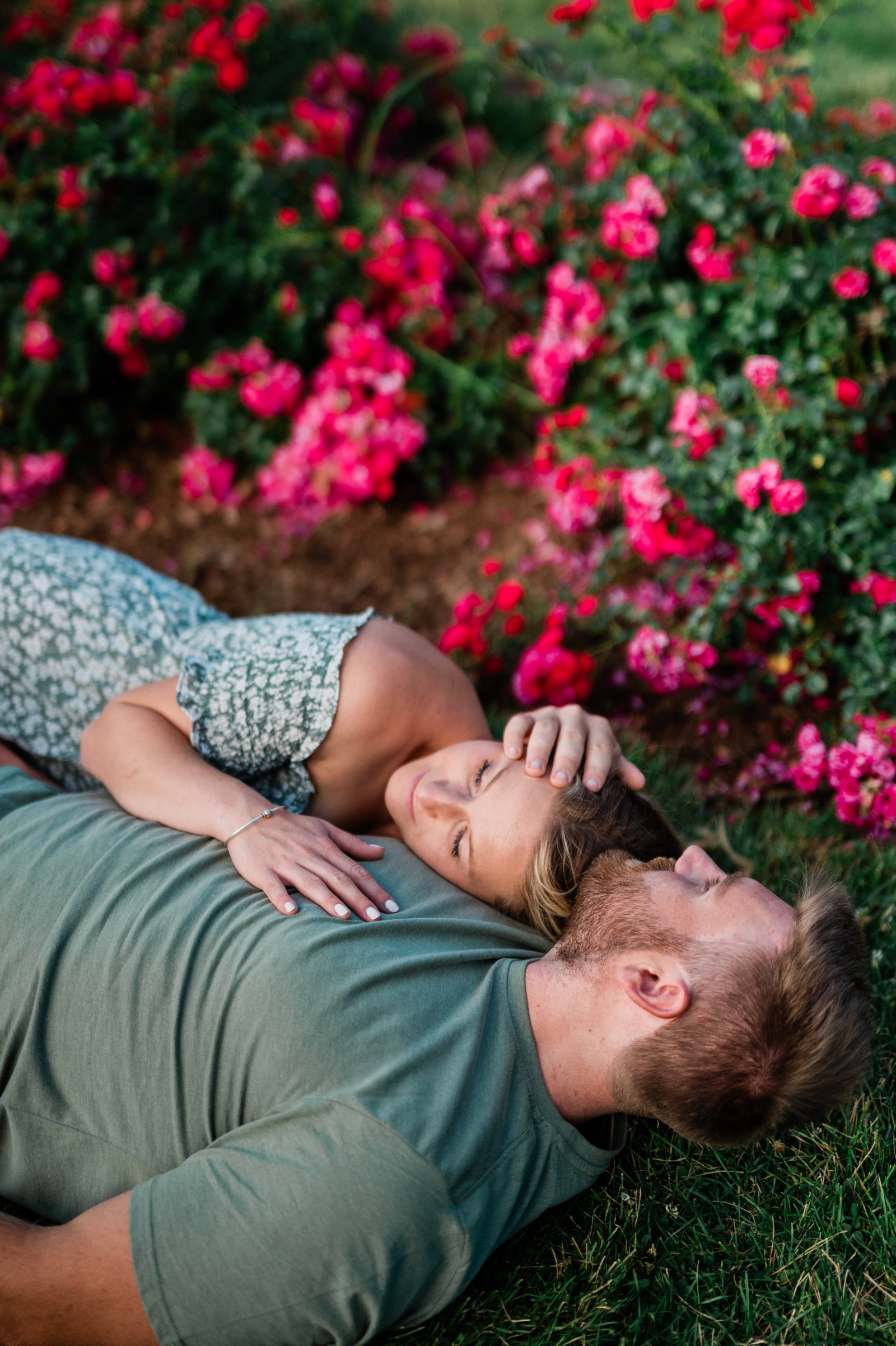 An engaged couple lays on the ground surrounded by roses at Elizabeth Park