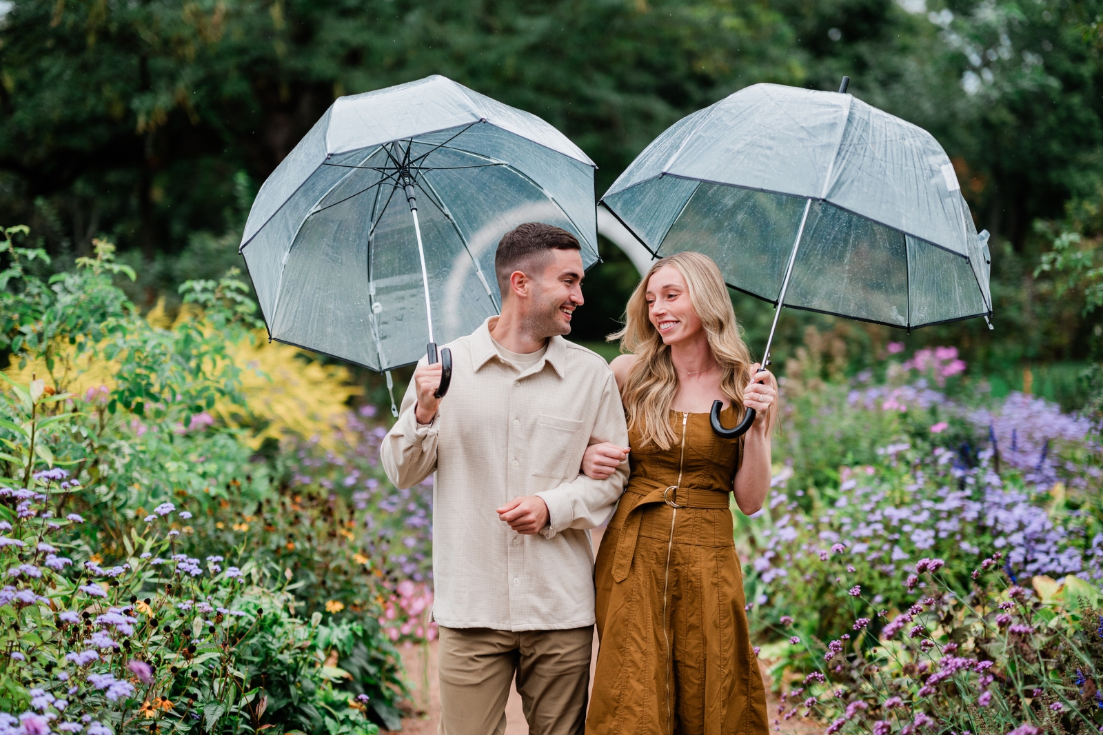An engaged couple walks through a wildflower garden in Weatherford, CT holding clear rain umbrellas