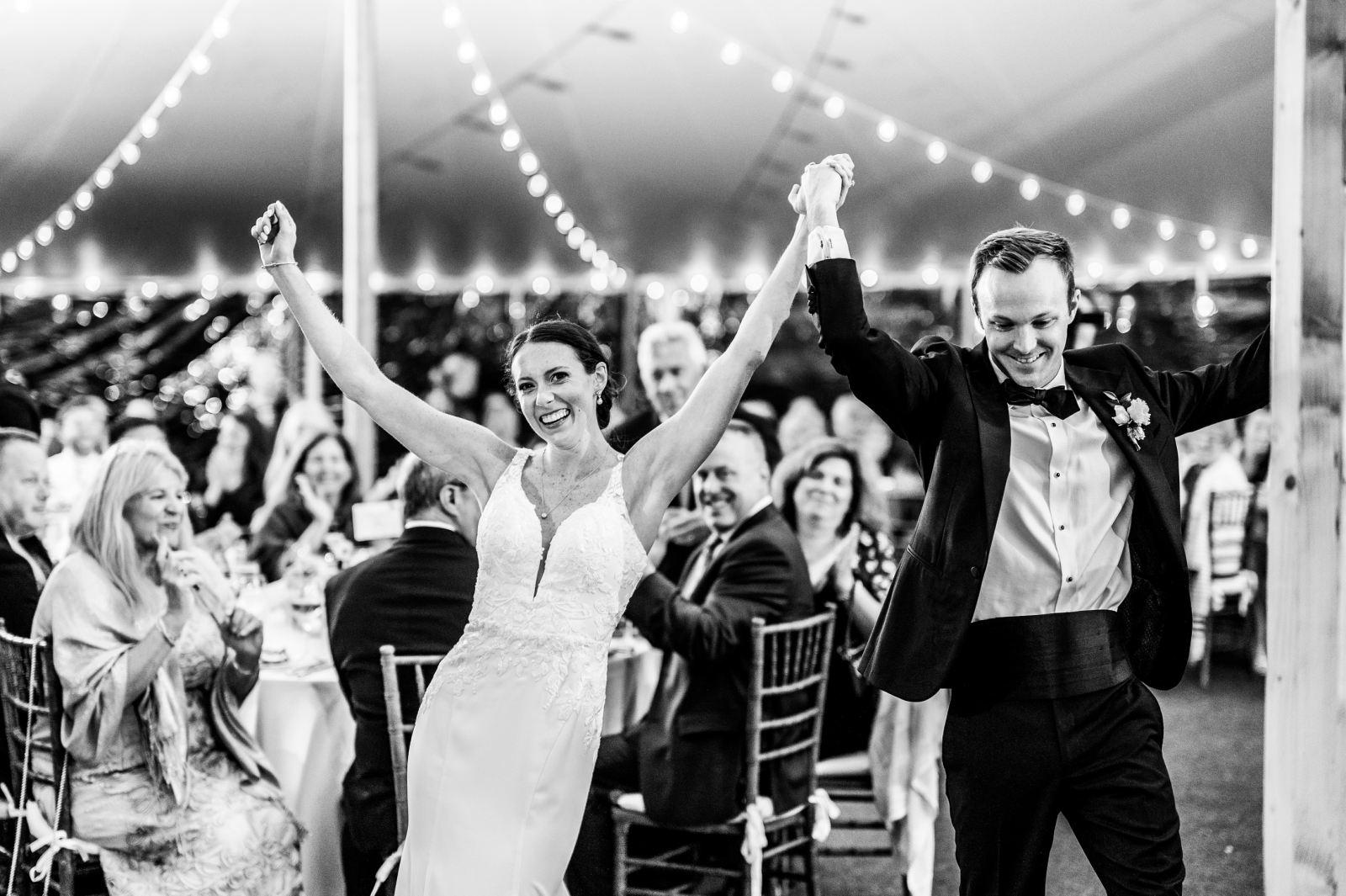 A black and white photo of a bride and groom entering their tented wedding reception while guests cheer 