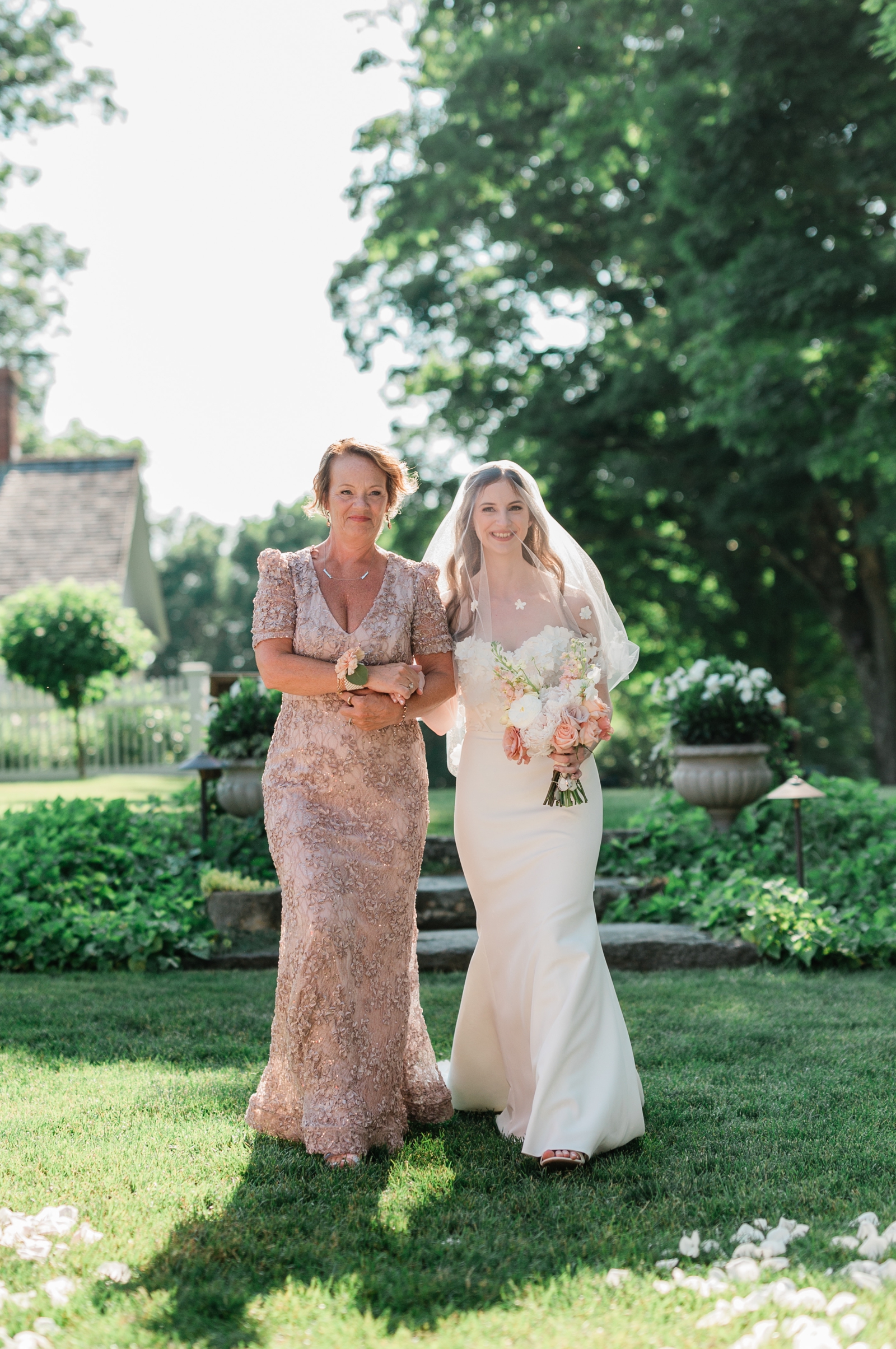The mother of the bride walks the bride down the aisle. The bride's face is covered by a tulle veil with applique flowers