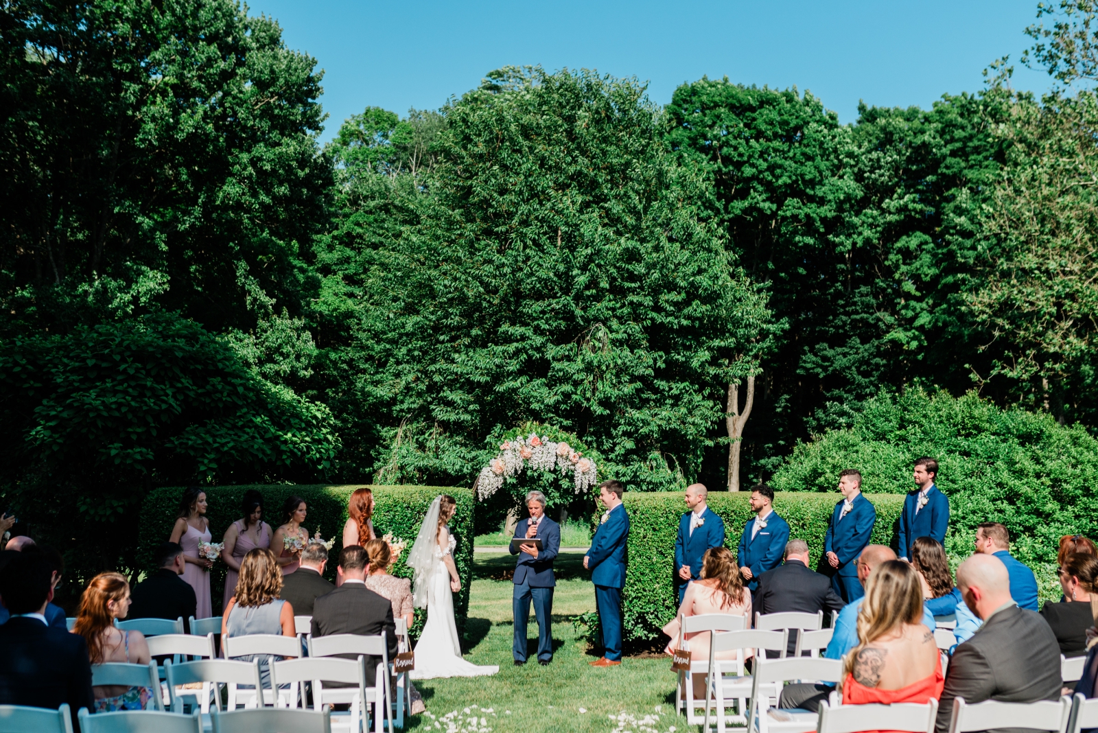 A bride and groom stand in front of a hedge with a floral arch at Smith Farm Gardens