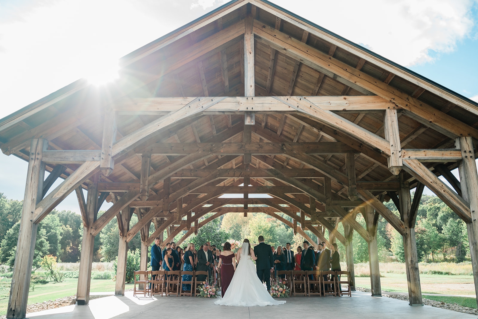 A bride walks down the aisle with her mother and father while the sun shines over the pavilion