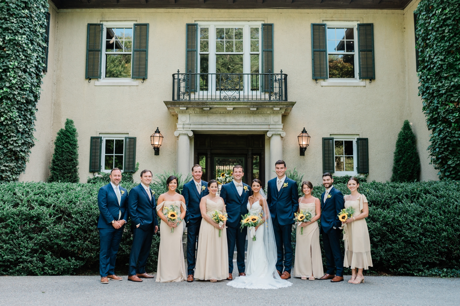 A wedding party dressed in navy suits and champagne dresses stands in front of Lord Thompson Manor