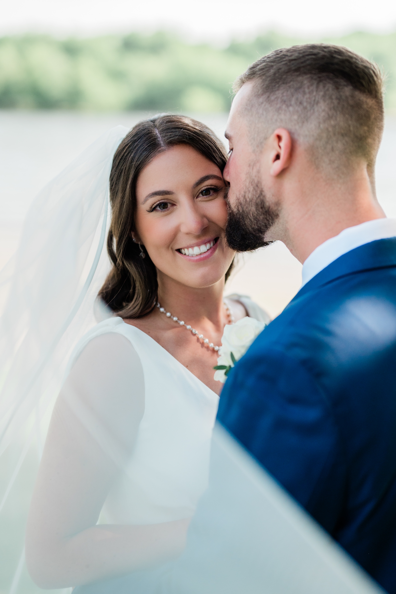 A groom kisses the bride on the cheek while she smiles at the camera