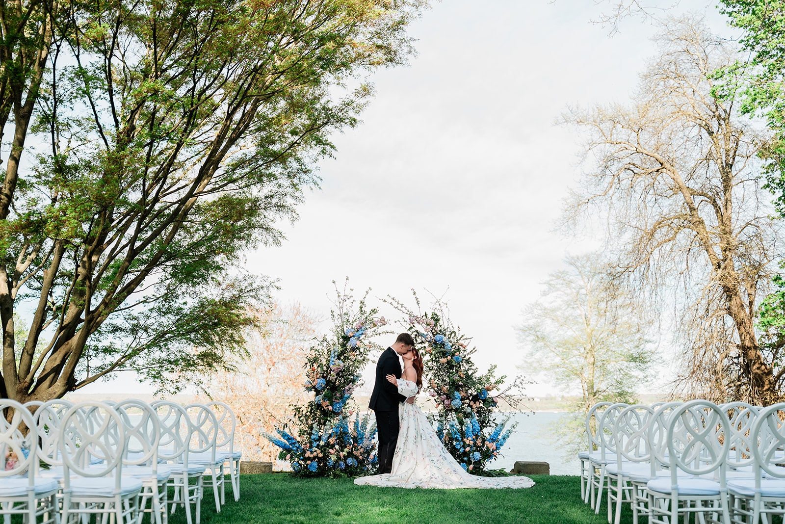 A bride and groom stand in front of a floral installation overlooking the Sakonnet River.
